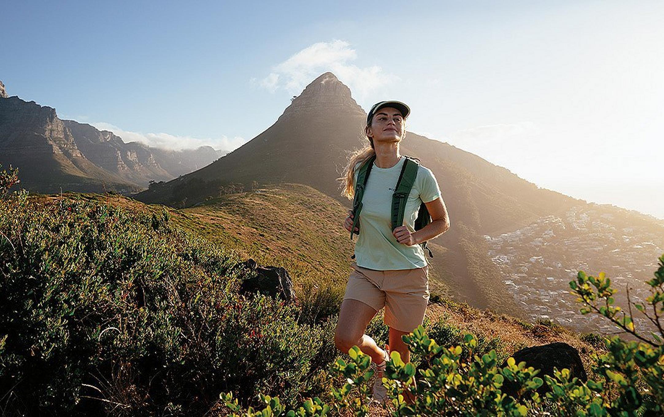 A female hiker with a backpack and sun-protective hat walks along a ridgeline after summiting a nearby peak.