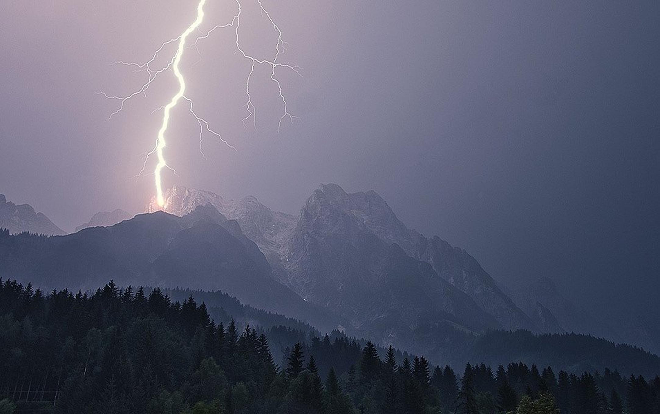 A high alpine peak is struck by a thick zig-zag of lightning that extends high above the mountain tops.