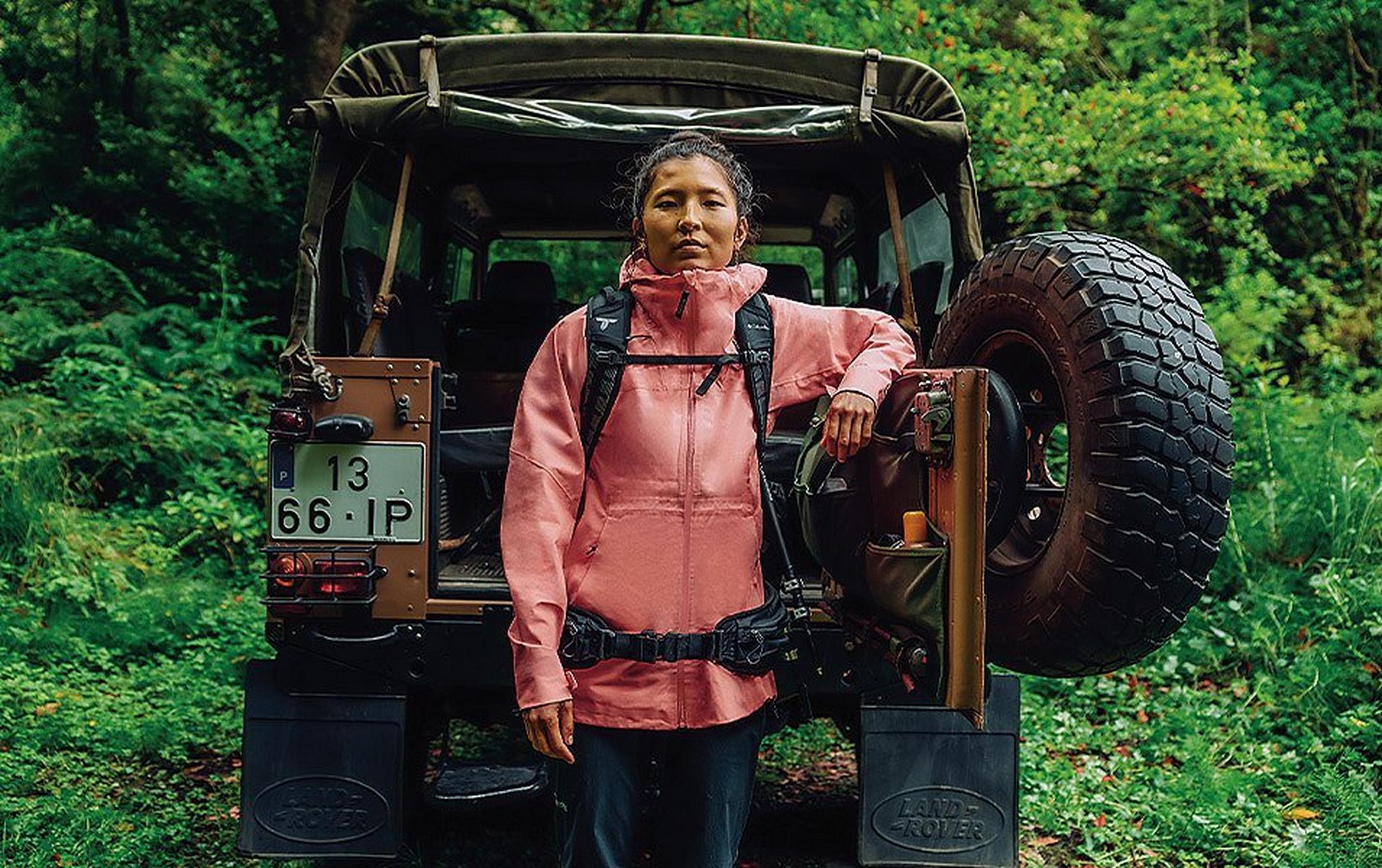 A woman, wearing a pack and a warm jacket,  leans against the back of a 4-wheel drive vehicle parked in a forest.