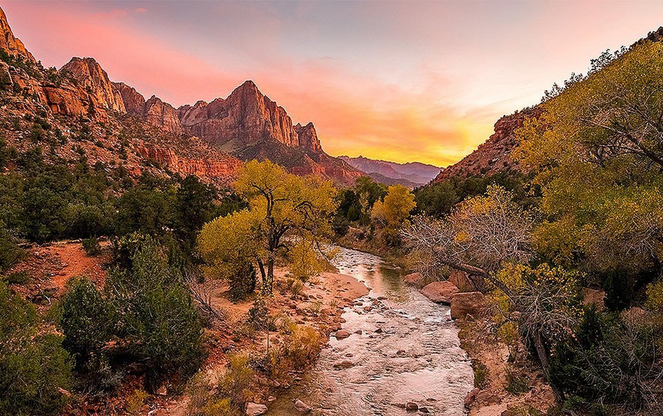 A dry, southwest landscape with banded sandstone cliffs in the background. A stream runs through the photo and the sky is turning yellow and orange.