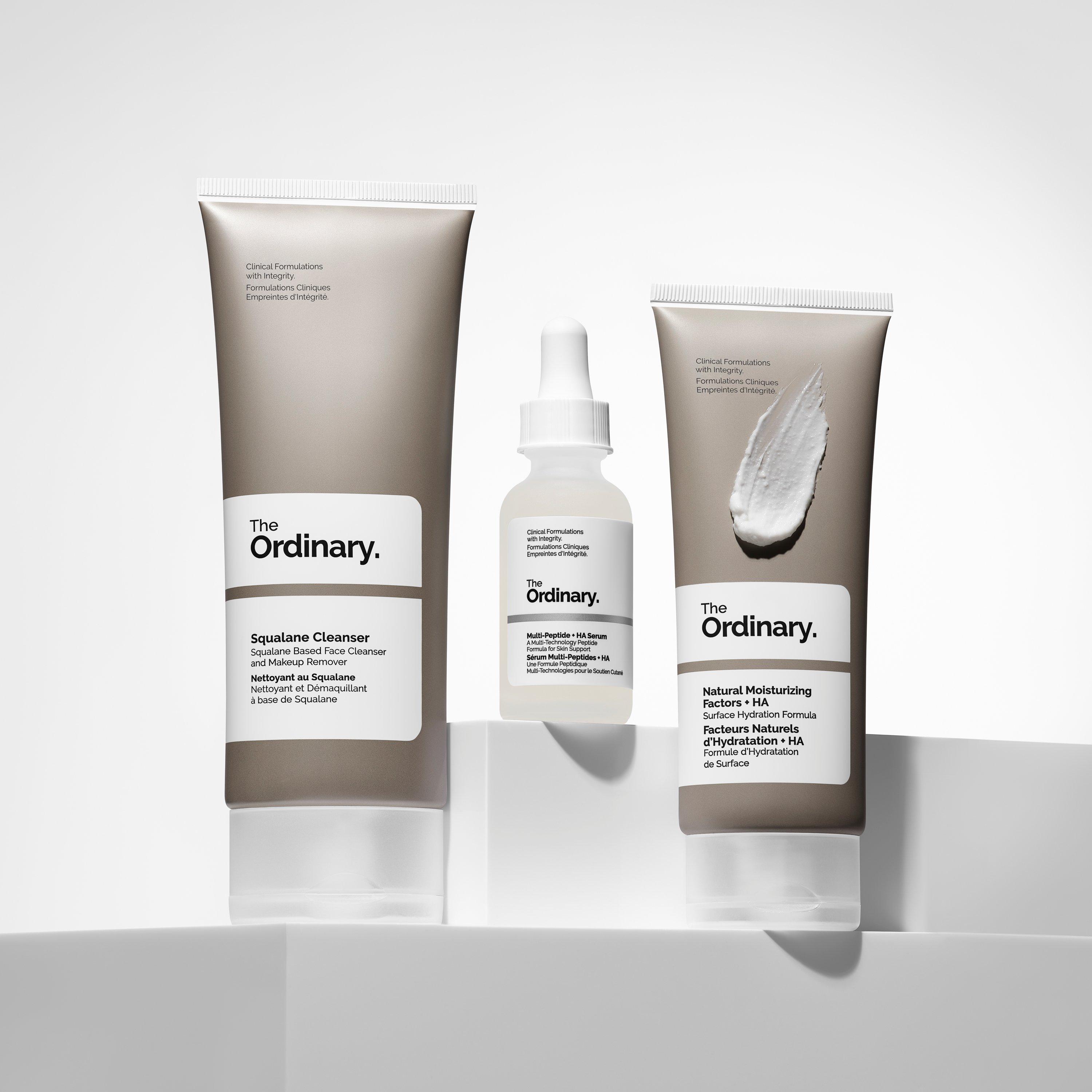 Three The Ordinary skincare products on a white surface: Squalane Cleanser in a tube, Multi-Peptide + HA Serum in a dropper bottle, and Natural Moisturizing Factors + HA in a tube with a swatch of cream.