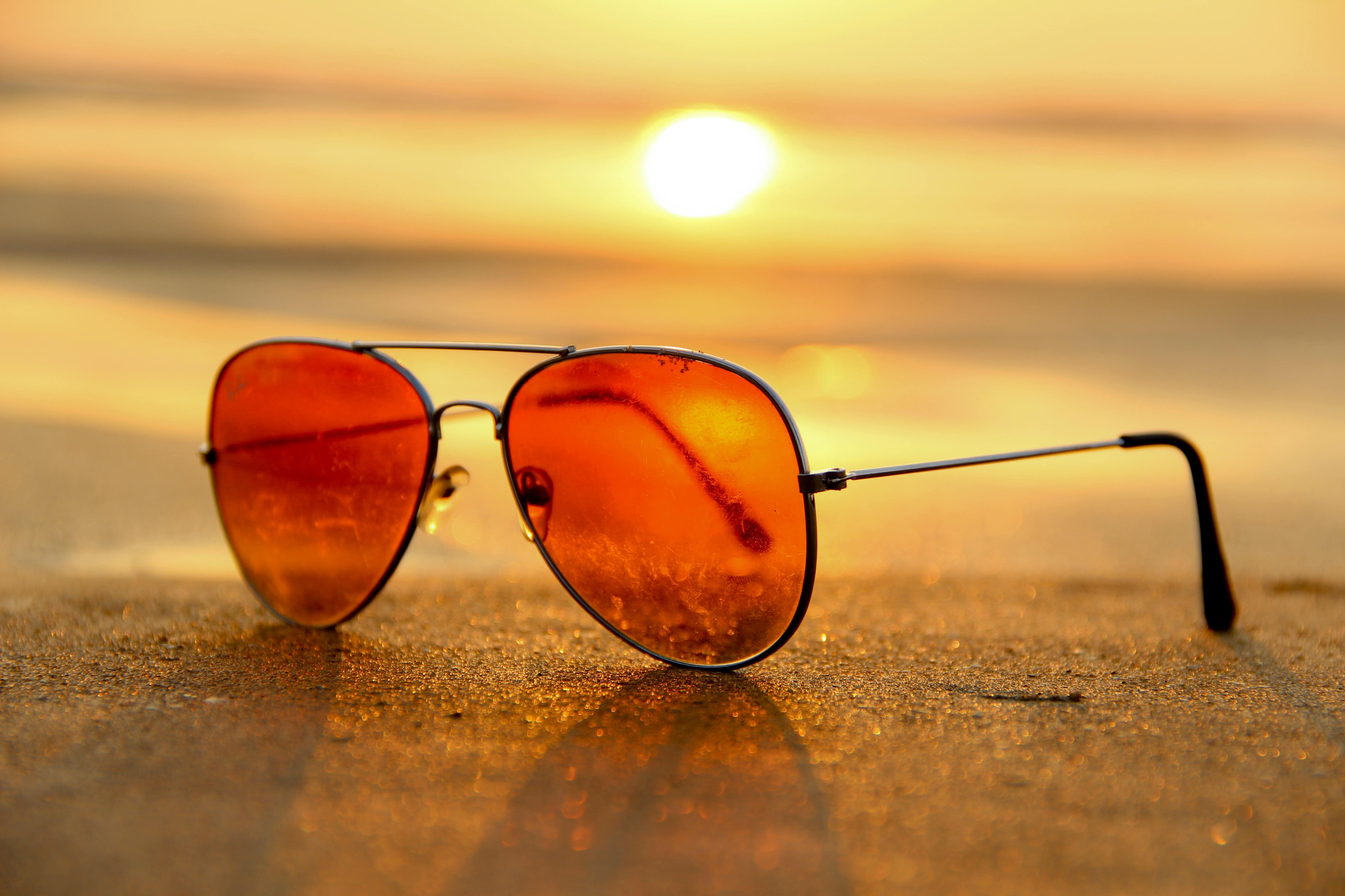 Aviator sunglasses with orange lenses placed on sandy beach at sunset.