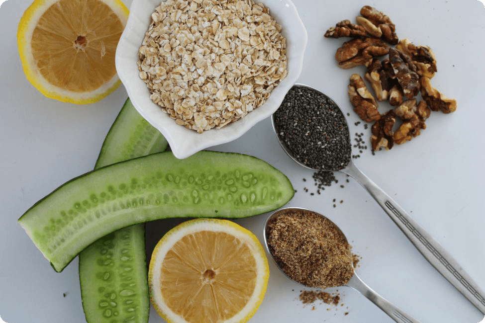 Slices of cucumber, lemon halves, rolled oats, walnuts, chia seeds, and ground flaxseed arranged on a white surface.