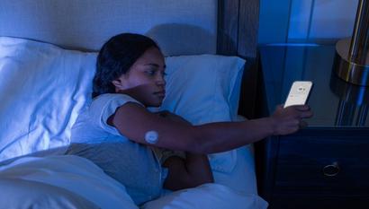Femme allongée dans son lit la nuit, regardant son smartphone équipé d'un capteur Dexcom G7 sur le haut du bras