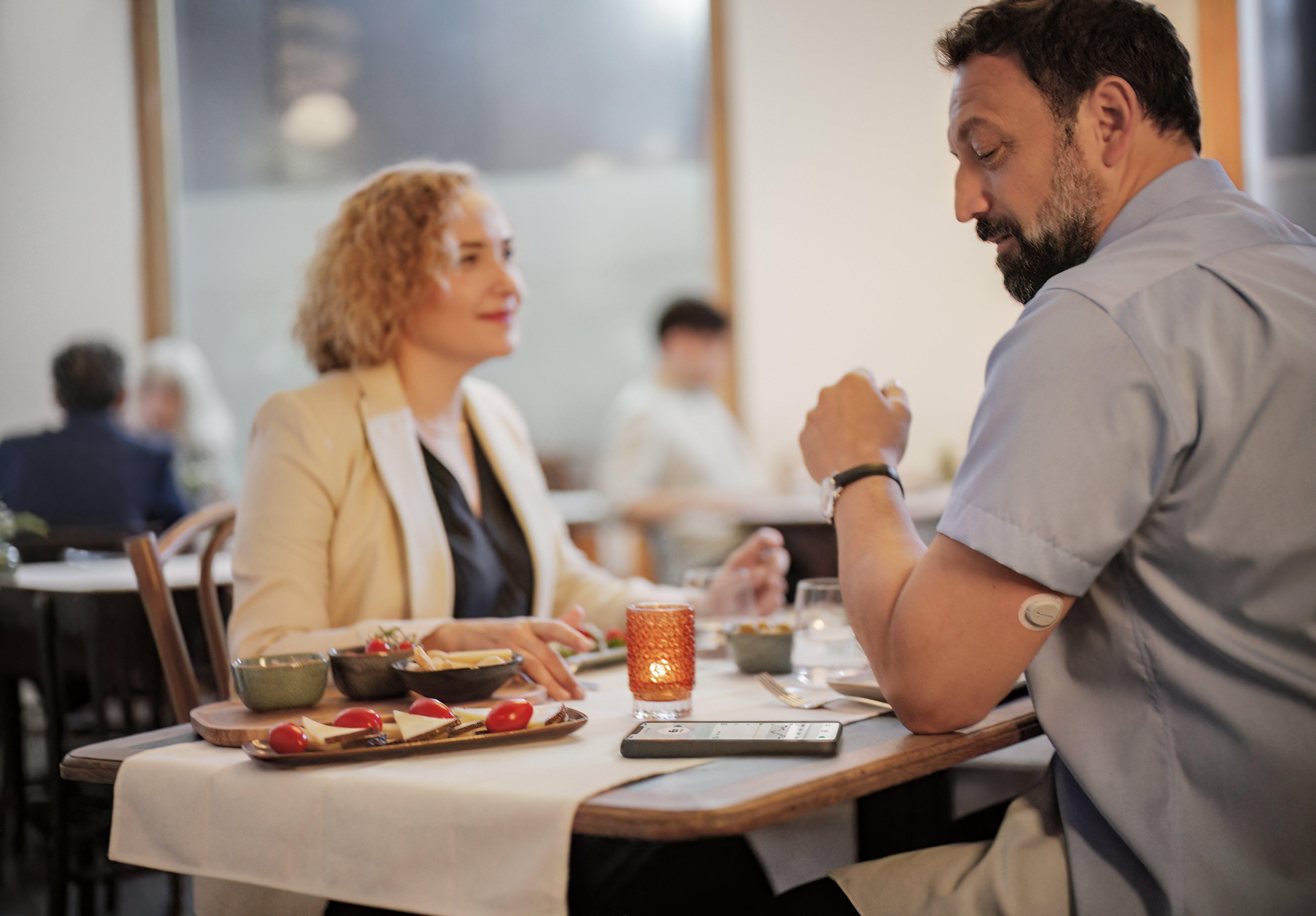 A man at a restaurant is wearing a Dexcom ONE+ device and checking the app