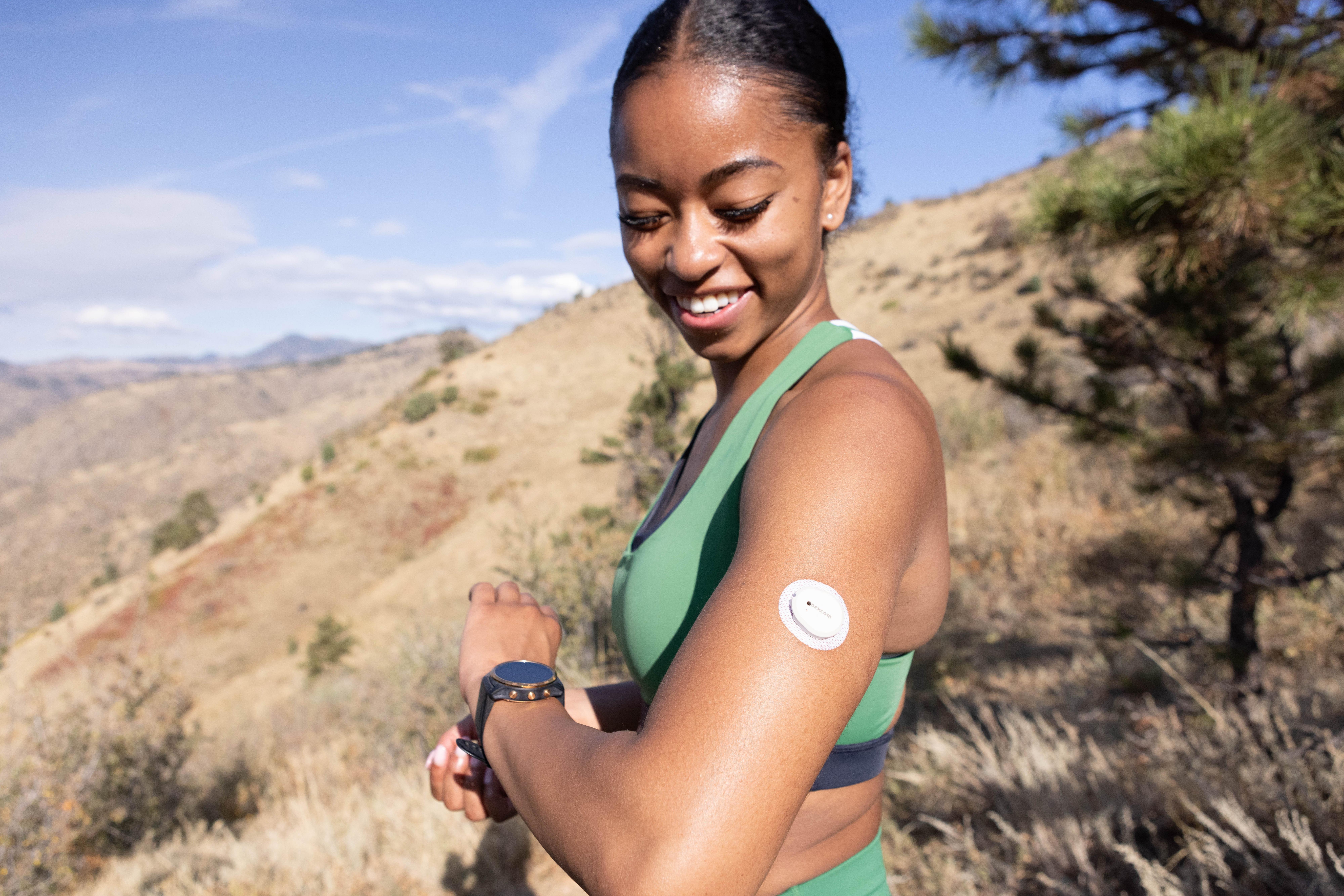 Lächelnde Frau draußen in den Bergen, die auf den Dexcom G7-Sensor an ihrem Oberarm schaut