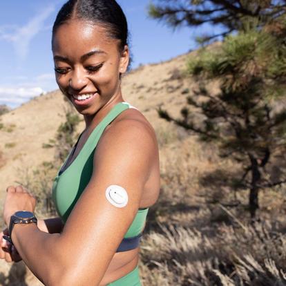 Lächelnde Frau draußen in den Bergen, die auf den Dexcom G7-Sensor an ihrem Oberarm schaut