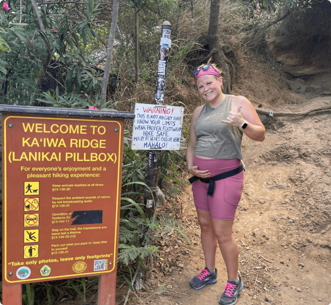 Devon holding her baby bump and holding up a "rock on" sign on KA'IWA Ridge trail