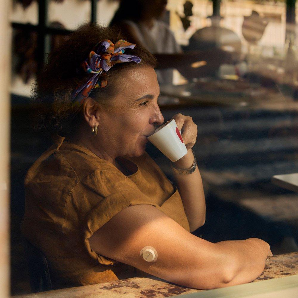 Eine Frau sitzt in einem Café, schaut zufrieden in die Ferne, während sie ihren Kaffee trinkt. Am Arm trägt sie den Dexcom Flex Sensor, der ihr das Fingerstechen erspart. 