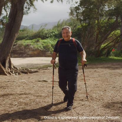 Older gentleman wearing a red backpack and holding two hiking sticks walking near trees