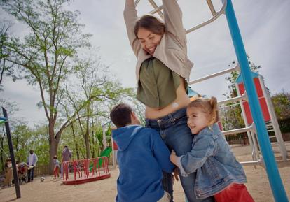 Mujer con un sensor Dexcom One+ en el abdomen jugando en un parque infantil con dos niños pequeños.