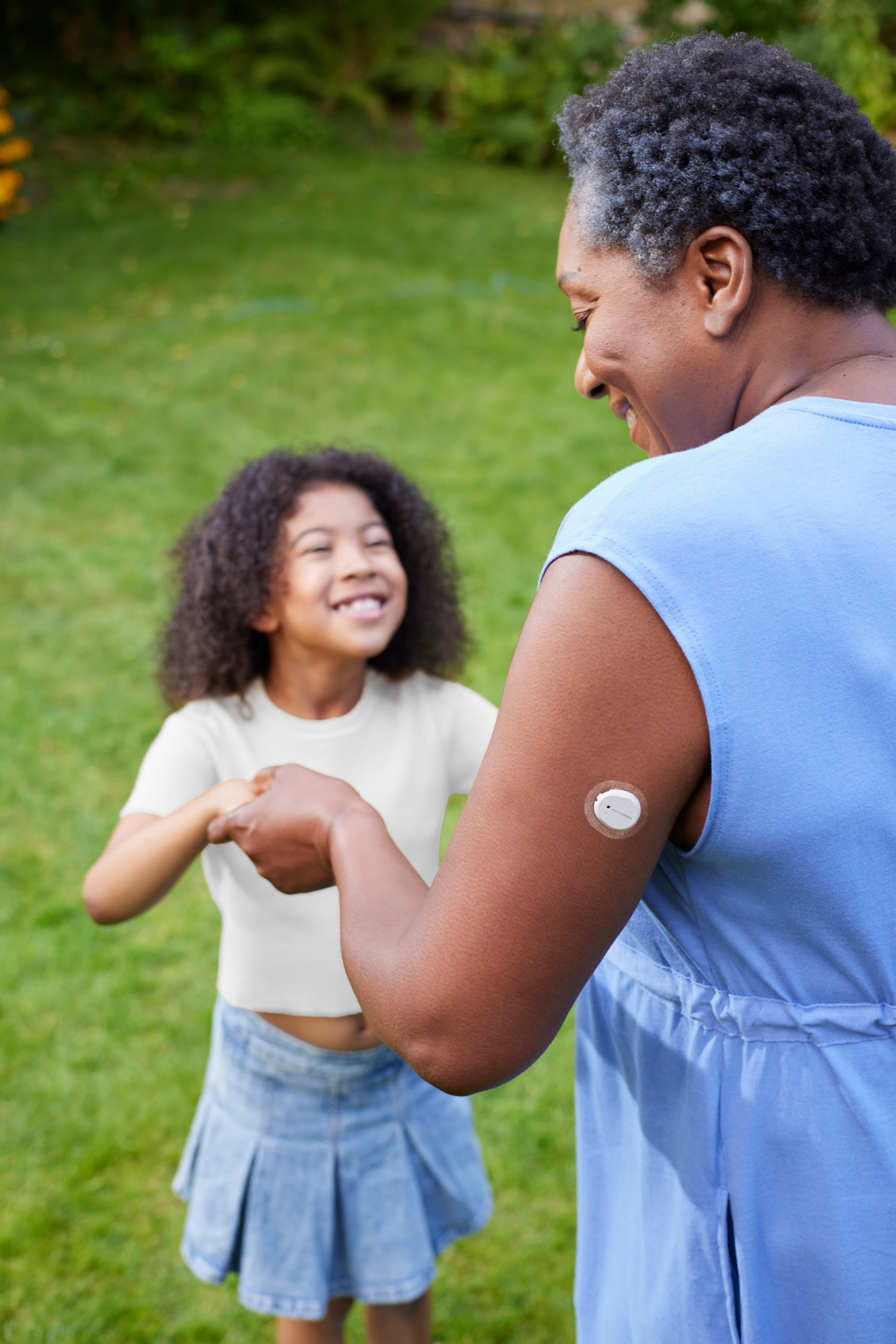 A woman wearing a Dexcom ONE+ on her arm is dancing with a child in the garden