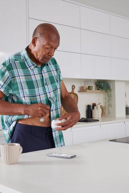 Older man standing in a kitchen applying a Dexcom ONE+ sensor on his abdomen