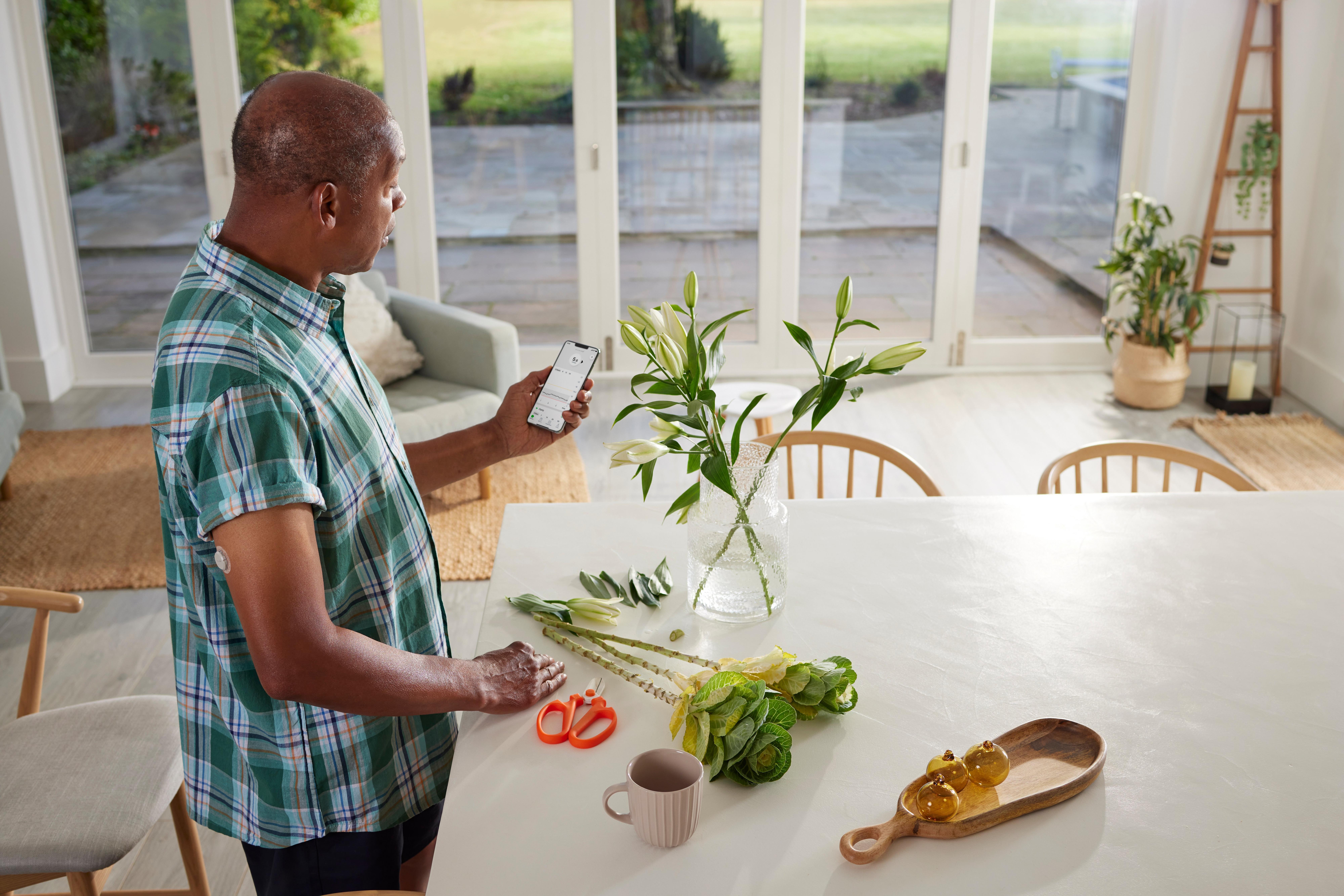 Man in keuken schikt bloemen en bekijkt de Dexcom ONE+ app op zijn smartphone