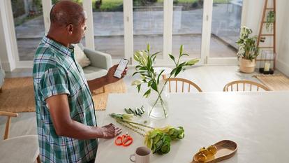 Mann in der Küche, der Blumen arrangiert und sich die Dexcom ONE+ App auf seinem Smartphone ansieht