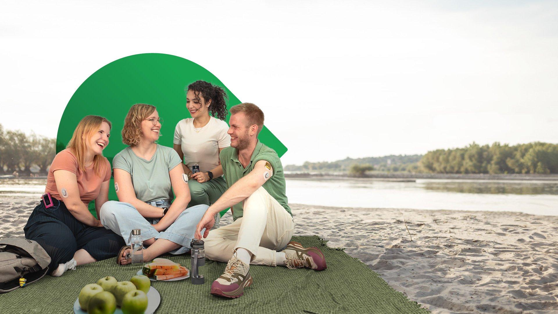 group of people smiling while having a picknick at a beach or sandy area with blurry water in the background. all the people displayed are wearing an insulin pump and a dexcom cgm.