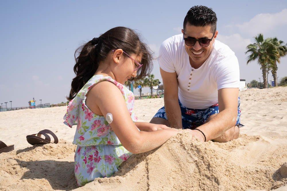 أب يلعب مع ابنته بجهاز D1+ على ذراعها في الرمال