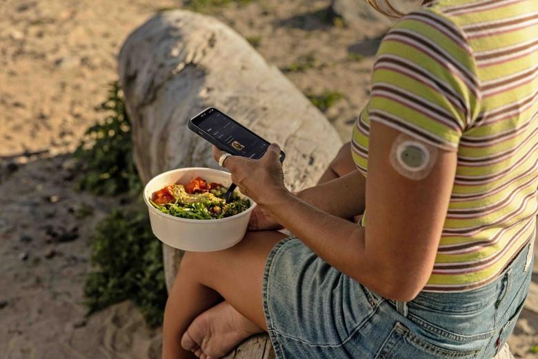A woman sitting outside on a rock holding a phone and a bowl of food