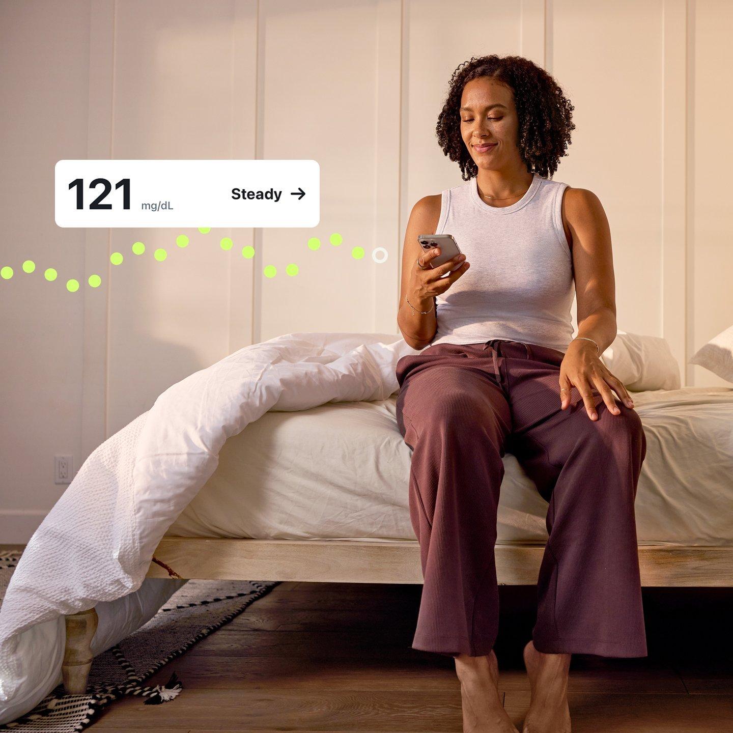 Woman sitting on bed, tracking her glucose levels on a smartphone