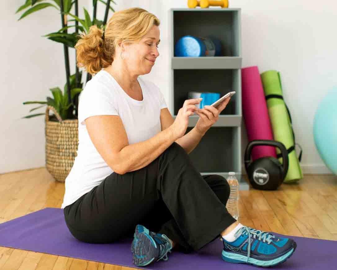 Sieviete sēž uz jogas paklājiņa un skatās uz tālruni