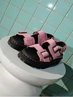 Sandalias rosas de lona con hebillas de latón y suela negra, sobre una peana blanca