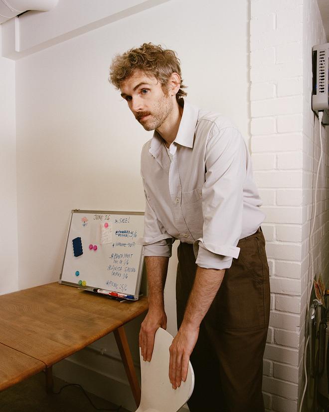 Person in a pale button-up shirt leans on a white chair beside a desk and small whiteboard