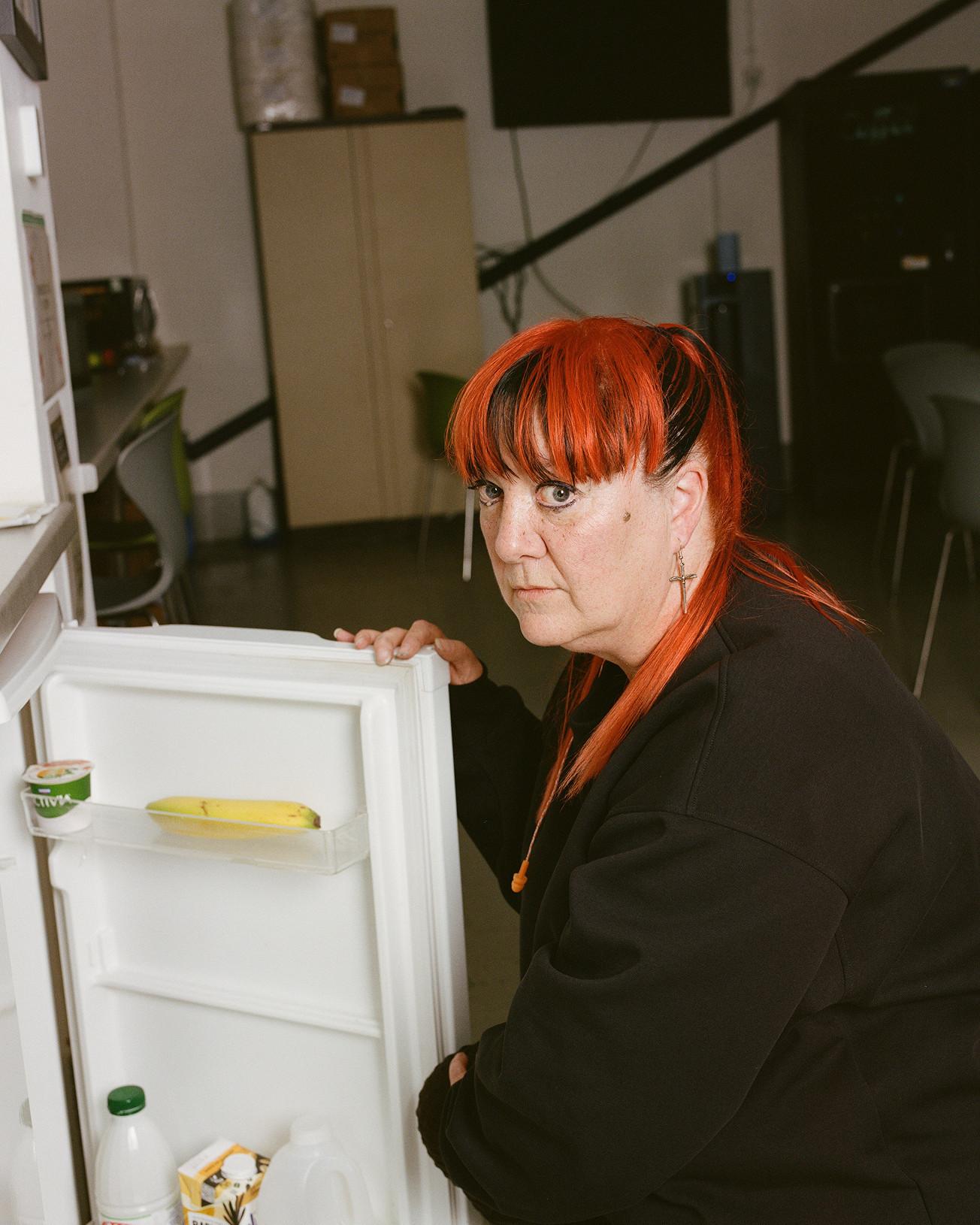 Persoon met feloranje haar in een zwarte hoodie hurkt bij een open koelkast
