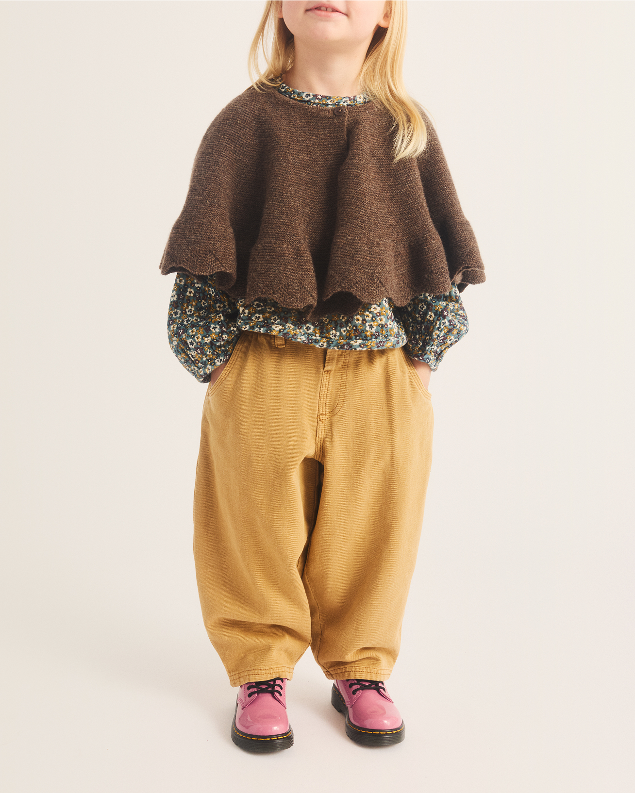 Child in pink lace-up Dr. Martens shoes, tan trousers and brown knit capelet