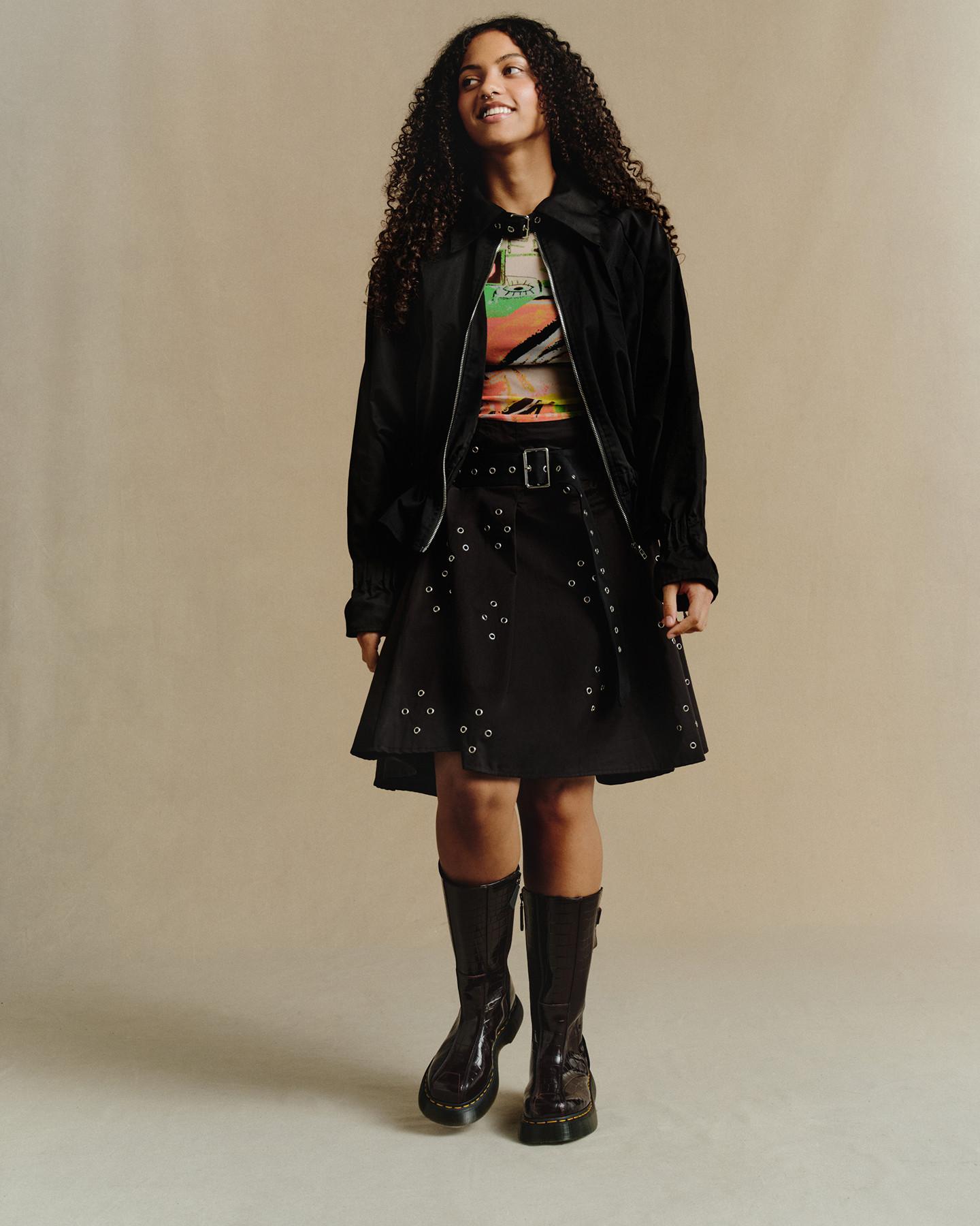 Model in shiny black knee-high boots, black mac and grommet skirt against a beige backdrop