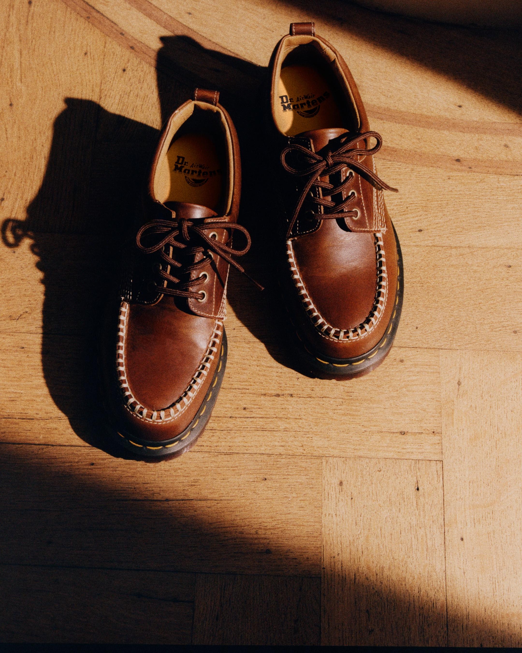 Kastanjebruine leren moc-toe schoenen met contrasterende stiksels, gefotografeerd in warm licht op een houten vloer