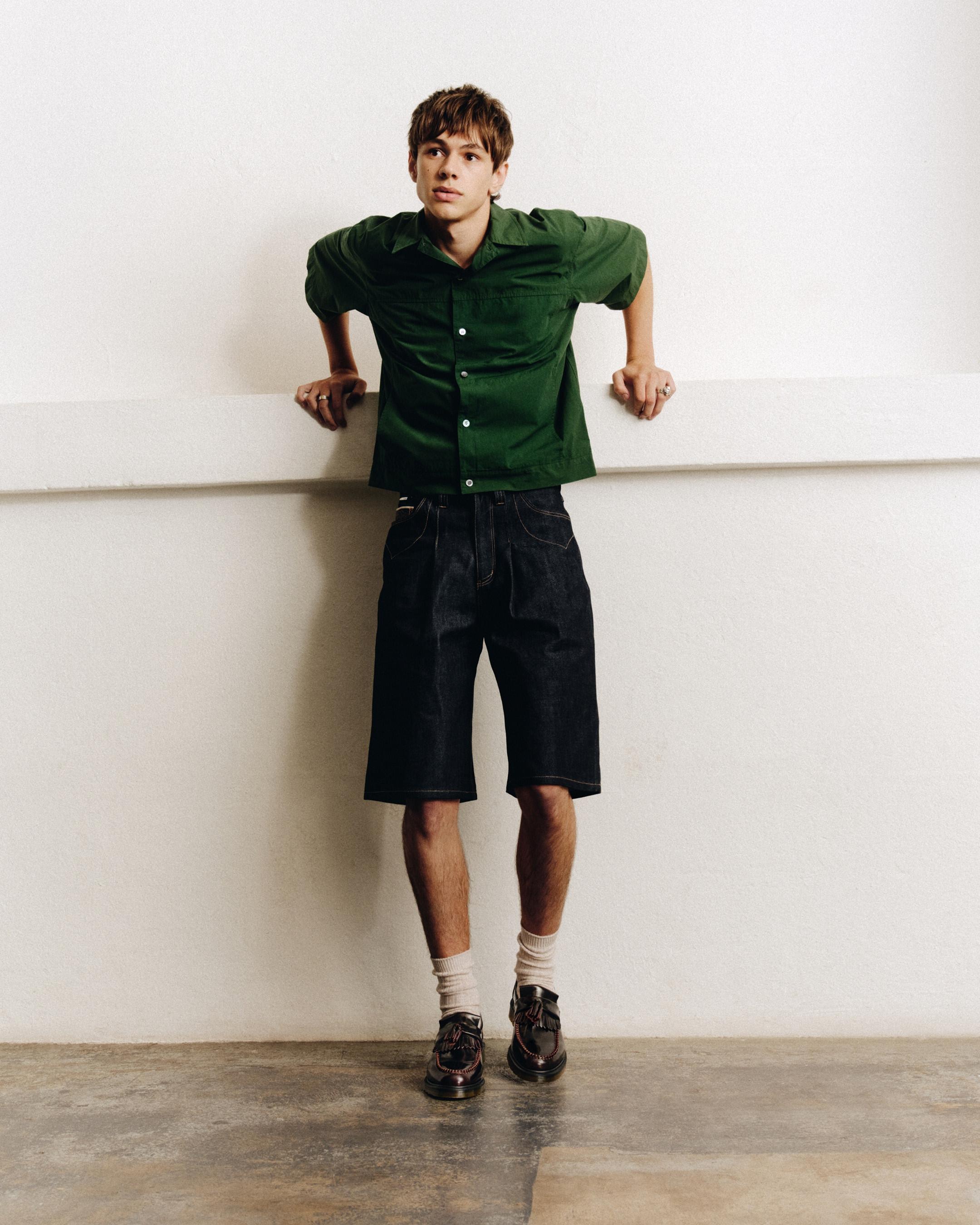 Person in green short-sleeve shirt and dark denim shorts, wearing black tassel loafers