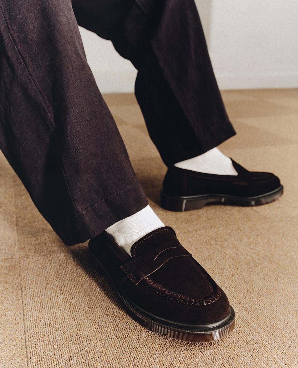Black suede penny loafers with white socks and dark trousers on a beige carpet