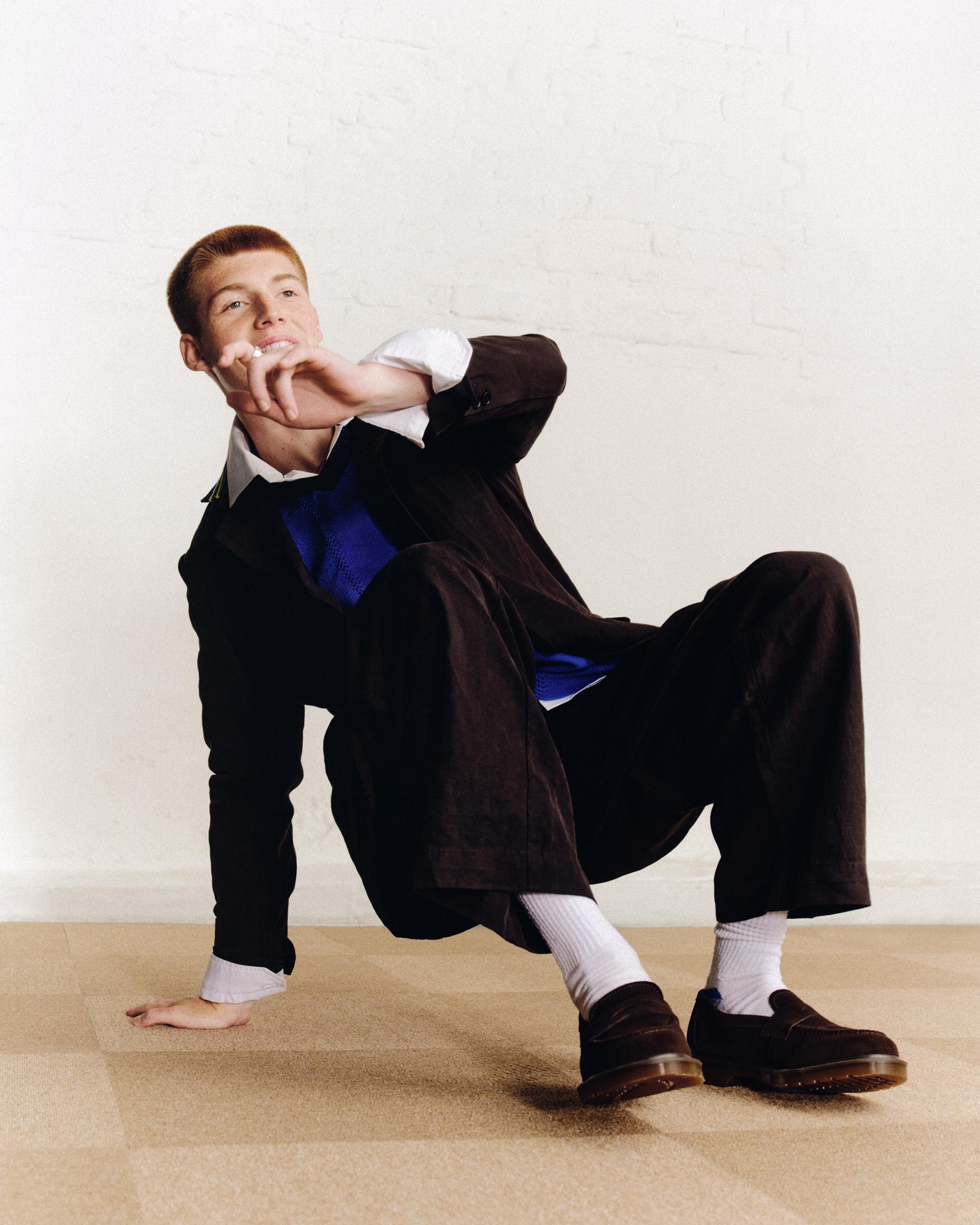 Person in a black suit and blue knit, lounging in dark leather loafers and white socks