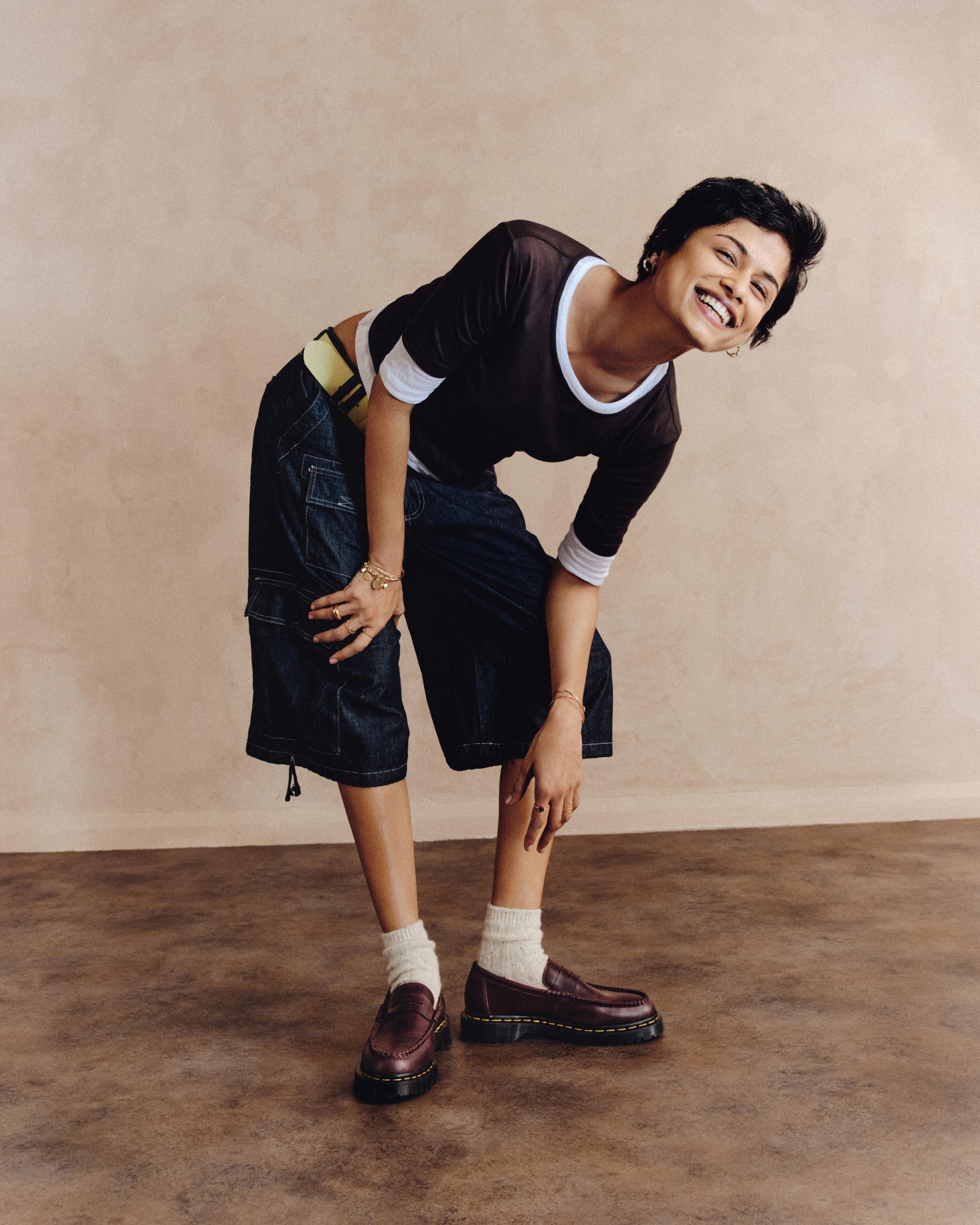 Person in burgundy leather loafers, white socks and dark denim shorts, leaning forward smiling