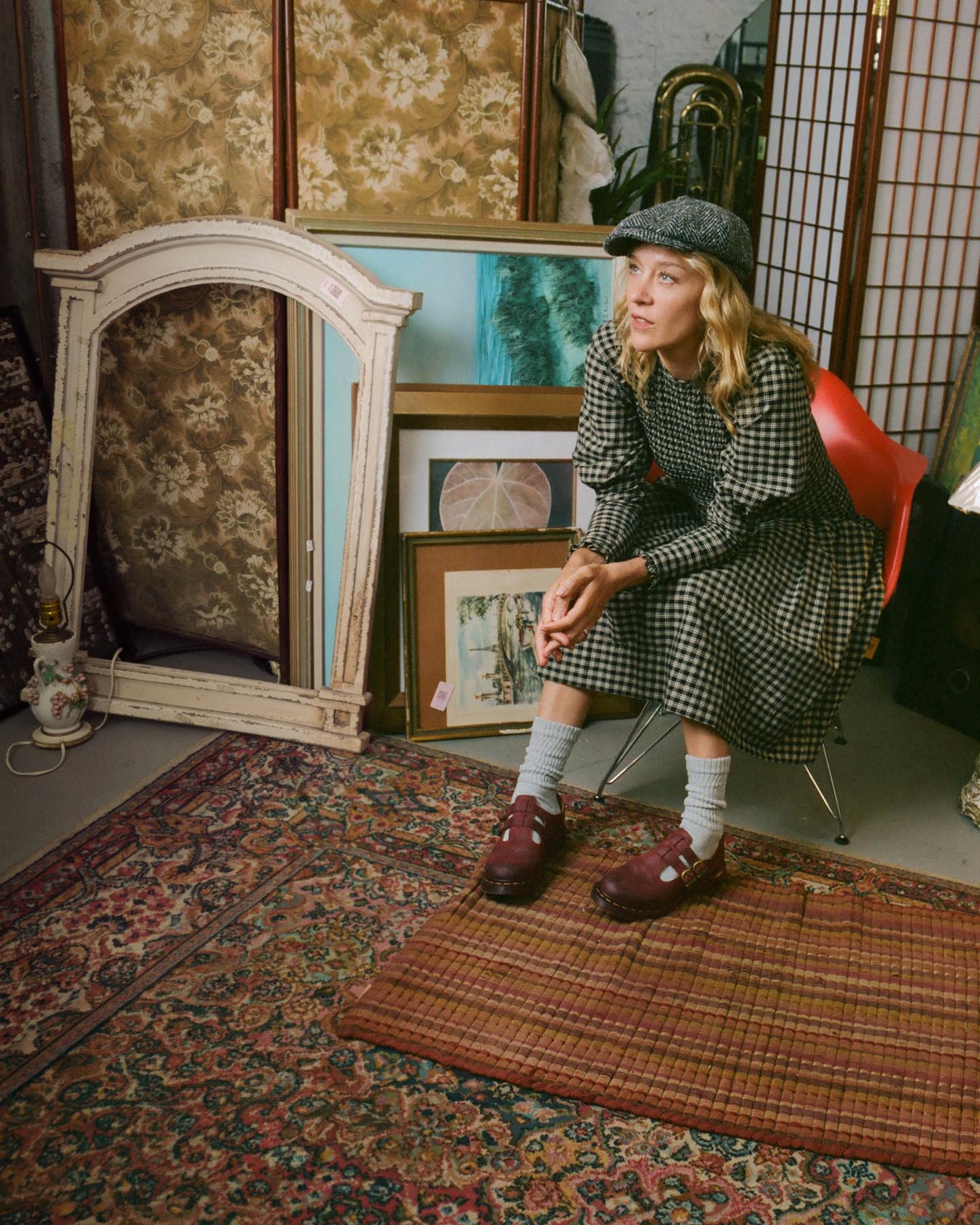 Person in a gingham dress and flat cap, seated, wearing burgundy Mary Jane shoes
