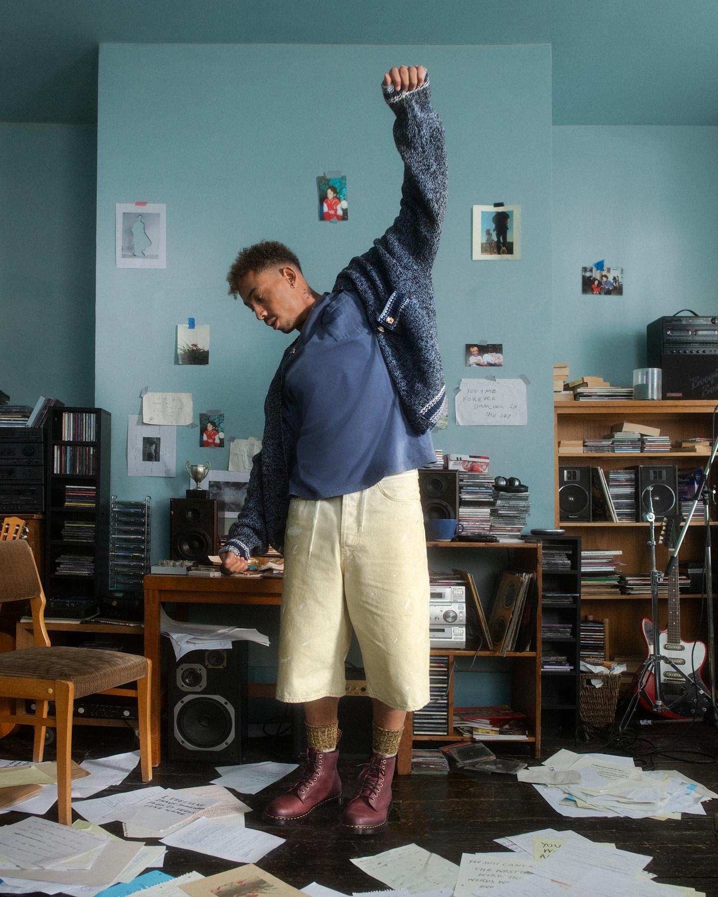 Person in burgundy lace-up boots, cream shorts and blue knit, arm raised in a messy music room