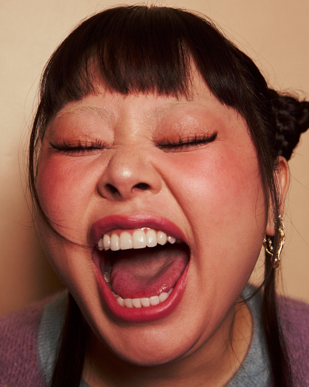 Close-up of a person laughing with eyes shut, bold pink lipstick and winged liner