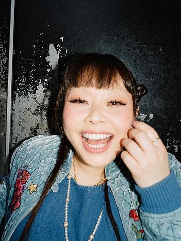 Smiling person in a blue knit jumper and embroidered denim jacket, close-up against a scuffed wall