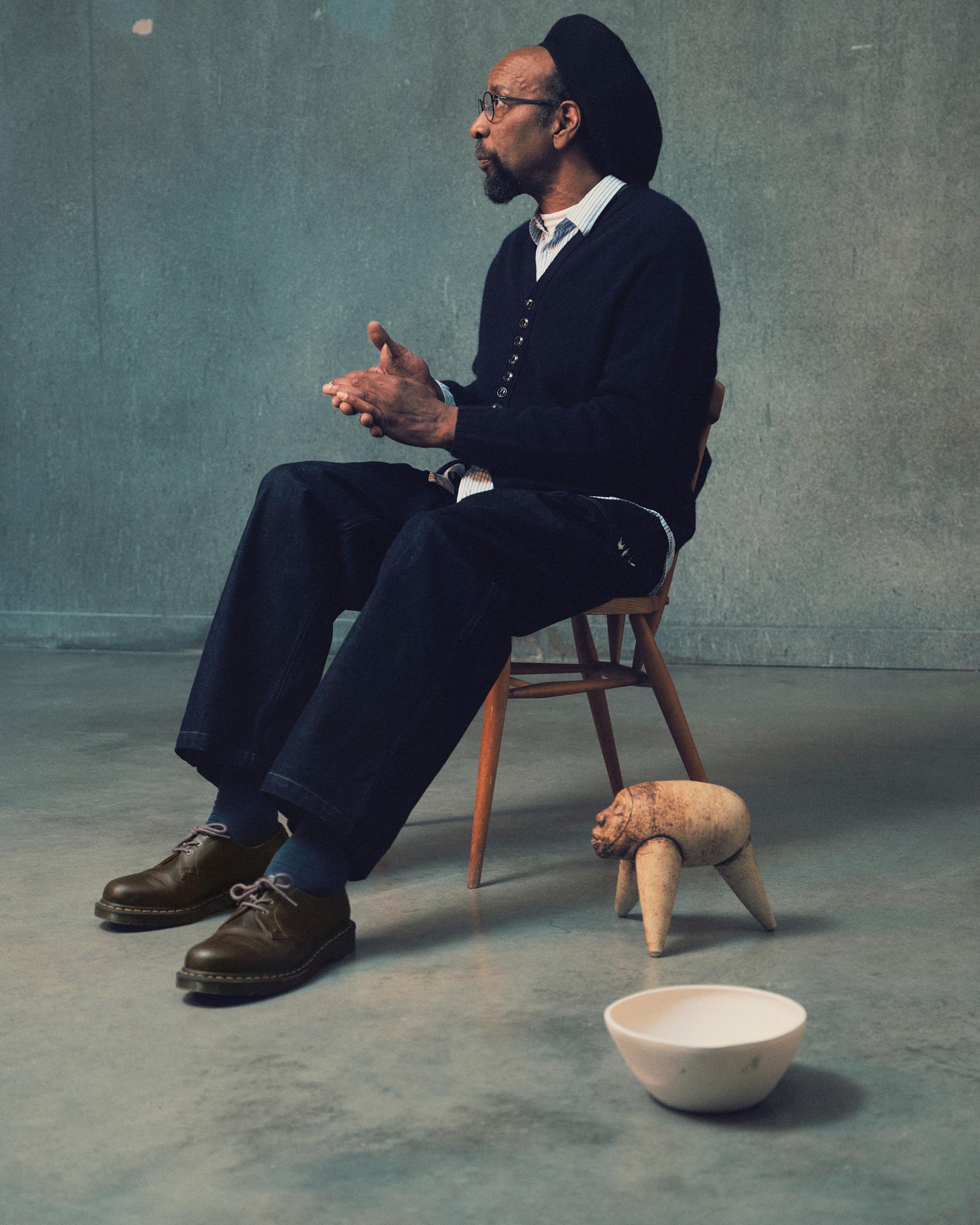Person in black cardigan and wide-brim hat, seated in worn leather shoes by a ceramic bowl