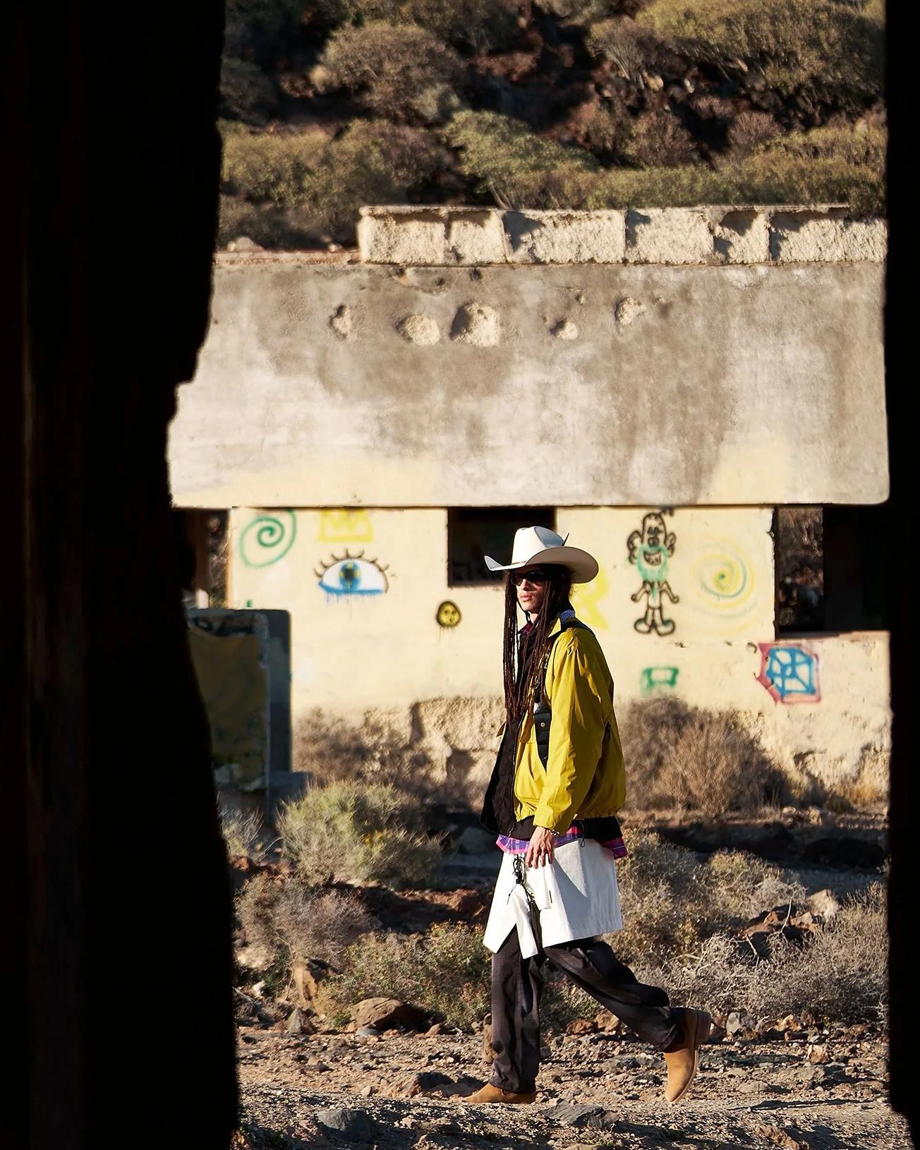 Person in white cowboy hat and yellow jacket walking past a graffiti-marked concrete bunker