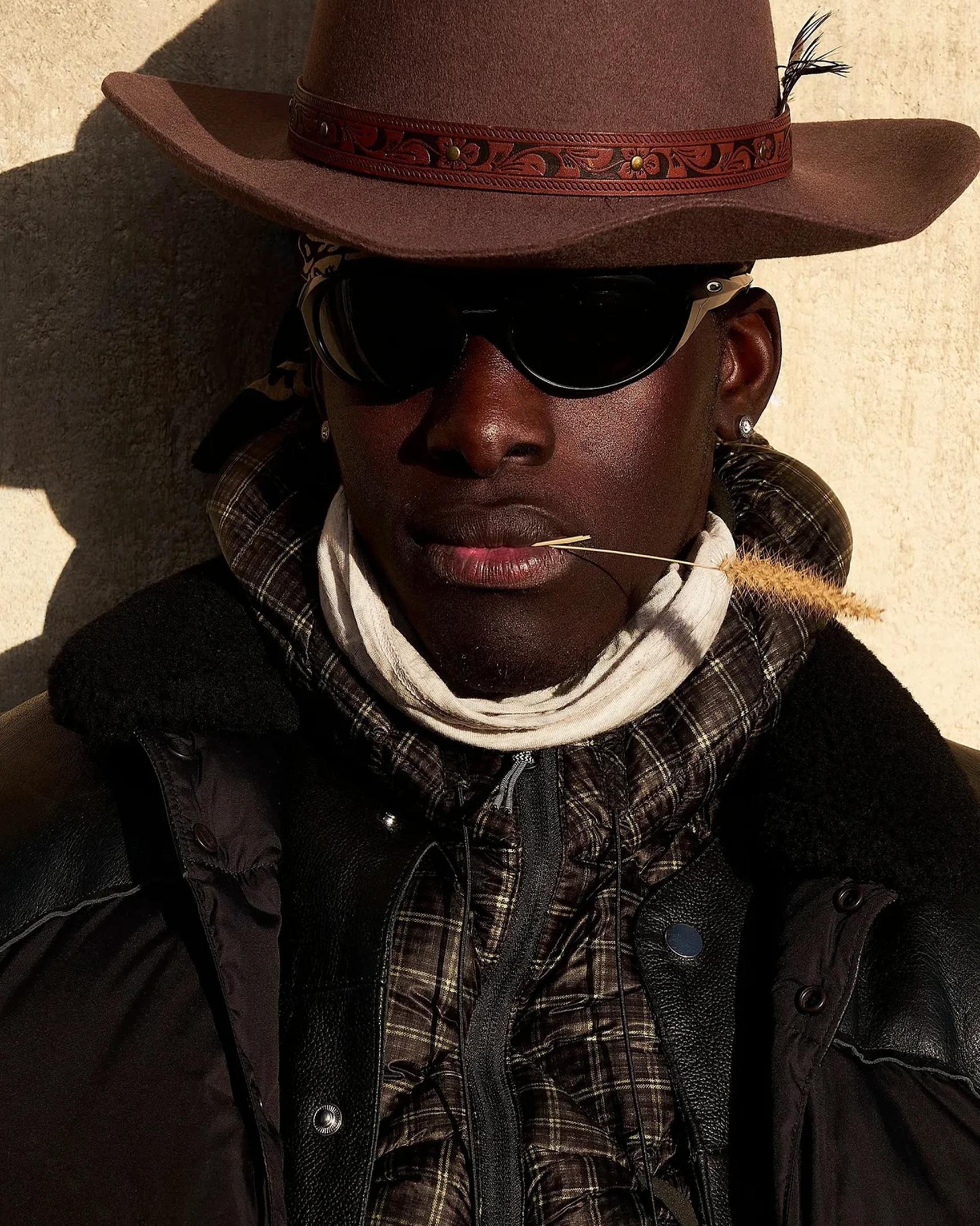 Person in a brown cowboy hat and sunglasses, dried grass stem in mouth, layered scarf and plaid jacket