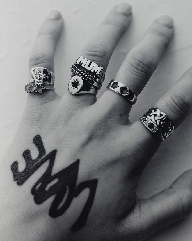 Nahaufnahme einer Hand mit silbernen Ringen, schwarze Kronen-Tätowierung am Handrücken