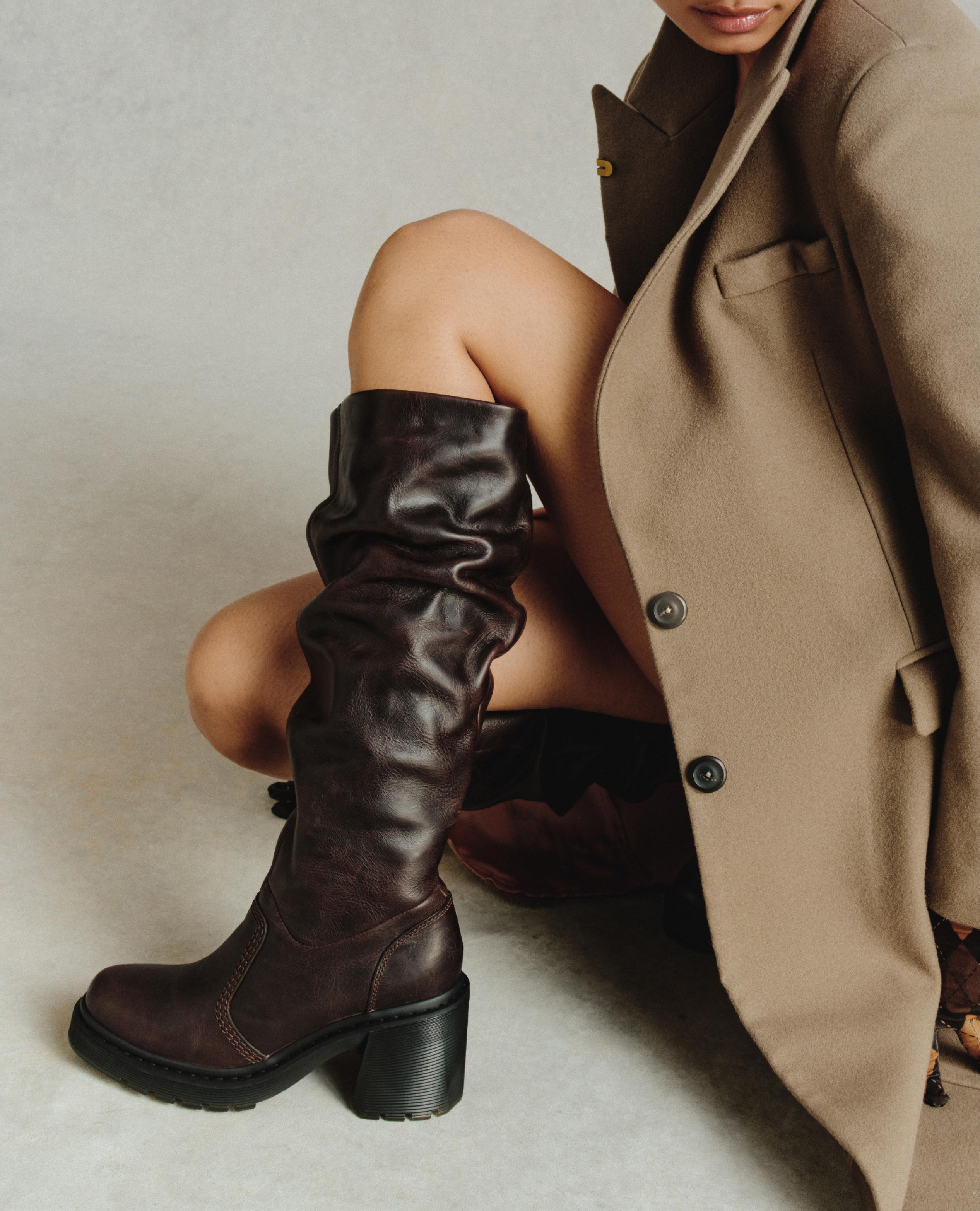 Modelo en cuclillas con abrigo camel y botas de cuero marrón oscuro, caña alta arrugada, tacón