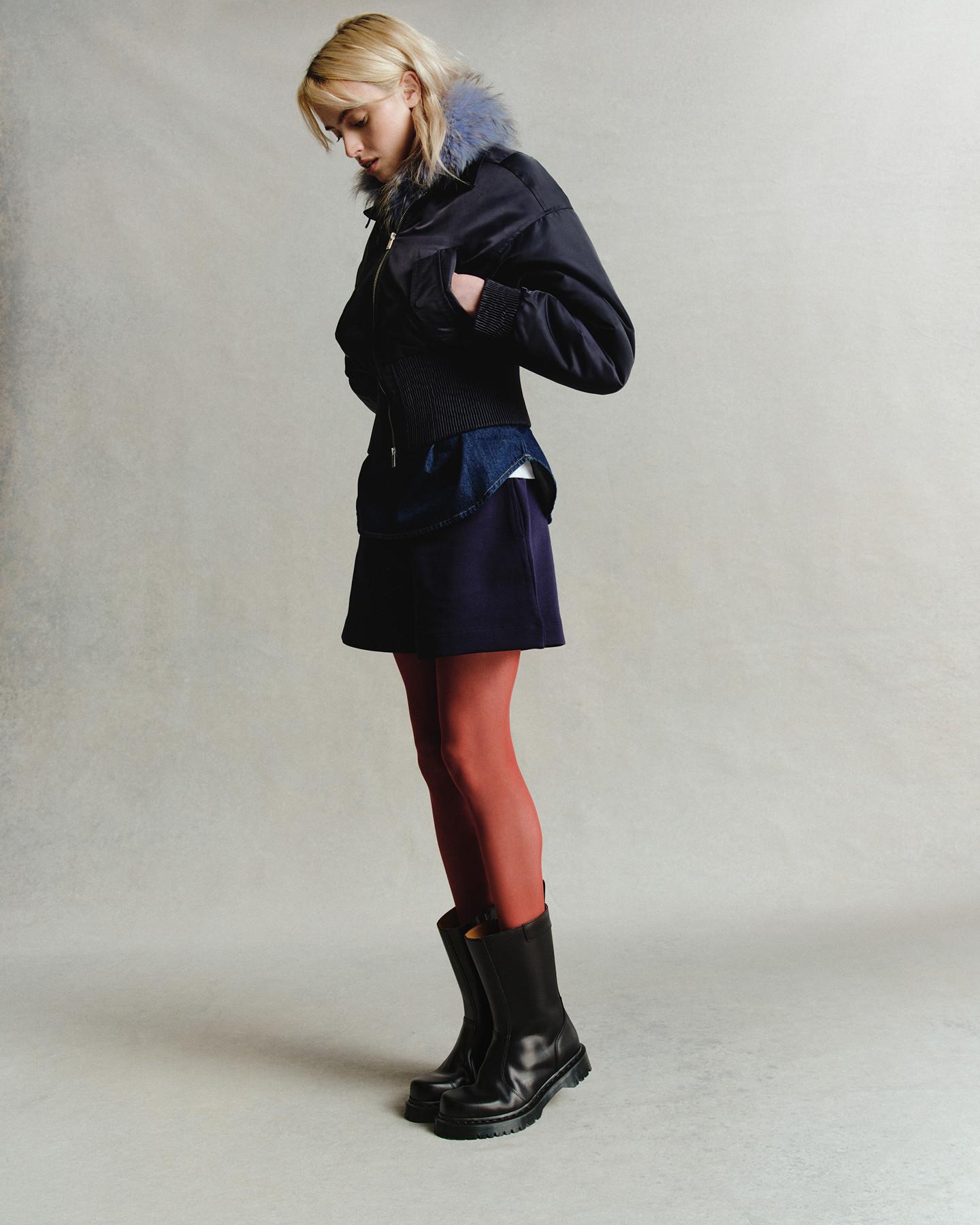 Persona con botas Chelsea de piel negra, falda azul marino y bomber con cuello de pelo sintético gris