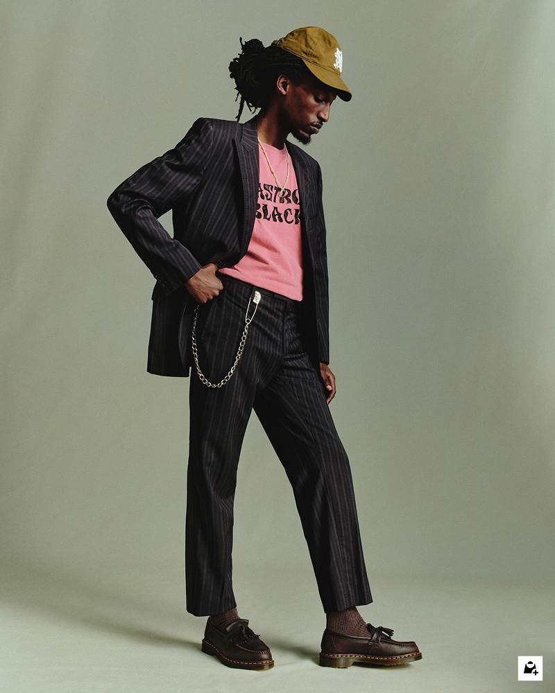 Person in black pinstripe suit and pink tee, wearing dark leather loafers