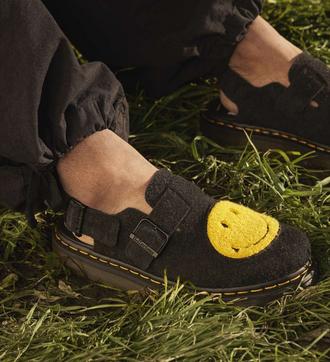 Zwarte schoenen met gele smiley, gedragen op gras. Dr. Martens logo op gesp.
