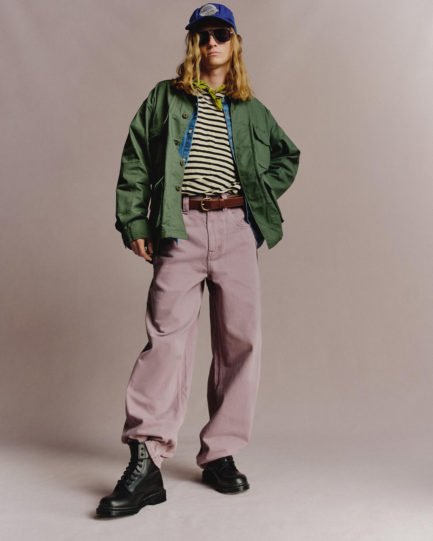 Person in black leather lace-up boots, green jacket and pale pink trousers against grey backdrop