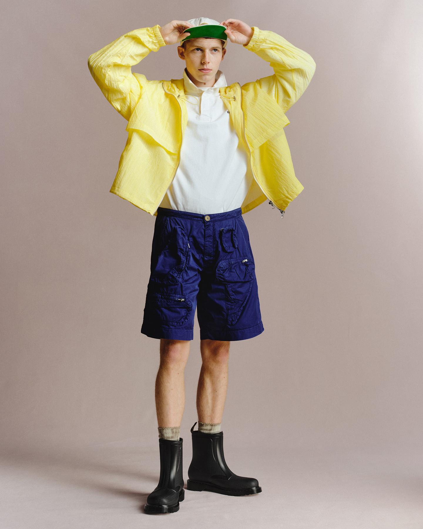 Person in black ankle boots, blue cargo shorts and a yellow jacket, adjusting a green cap