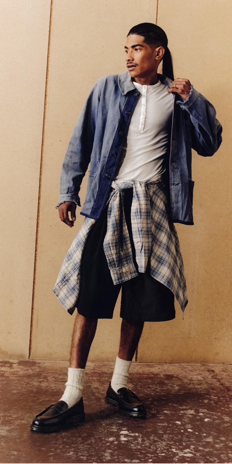 Person in black loafers, white socks and layered workwear with a plaid shirt tied at the waist