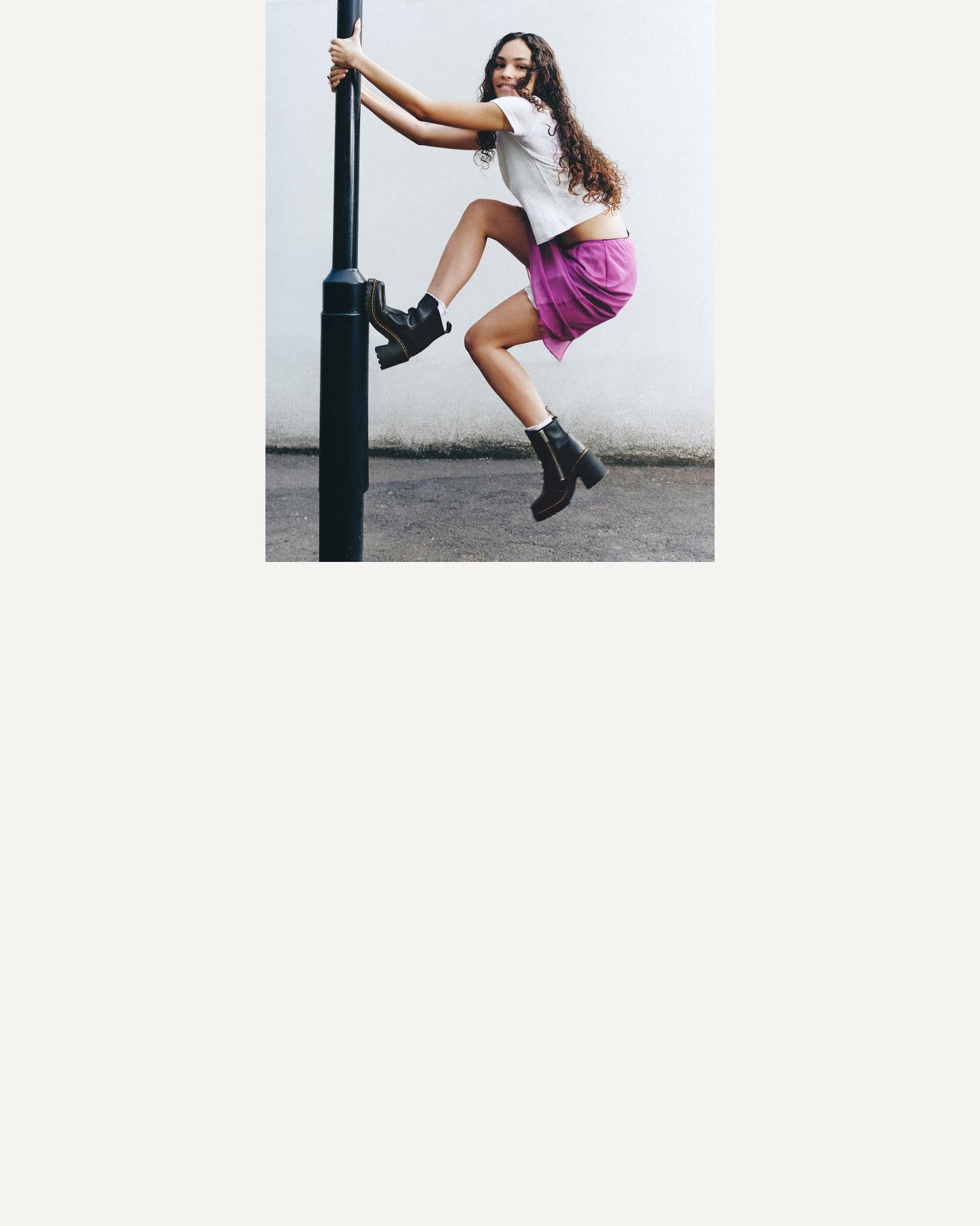 Person climbing a street pole in black leather ankle boots, white tee and purple skirt